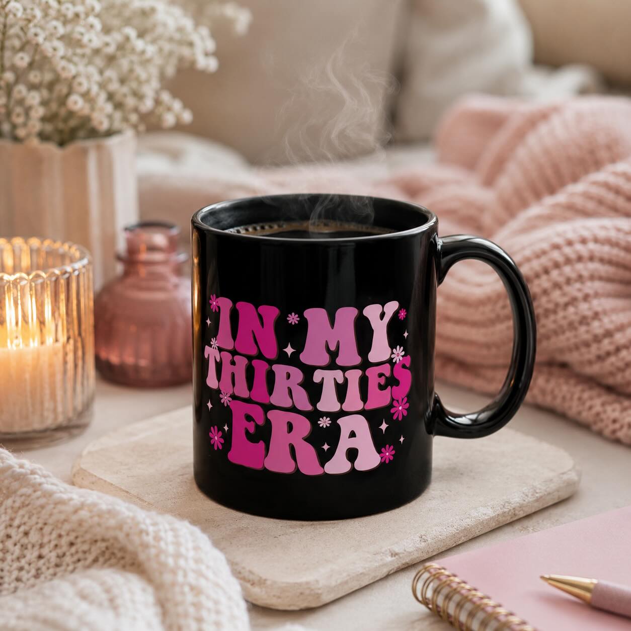 In My Thirties Era black mug styled with candle, dried flowers, and pink knit blankets